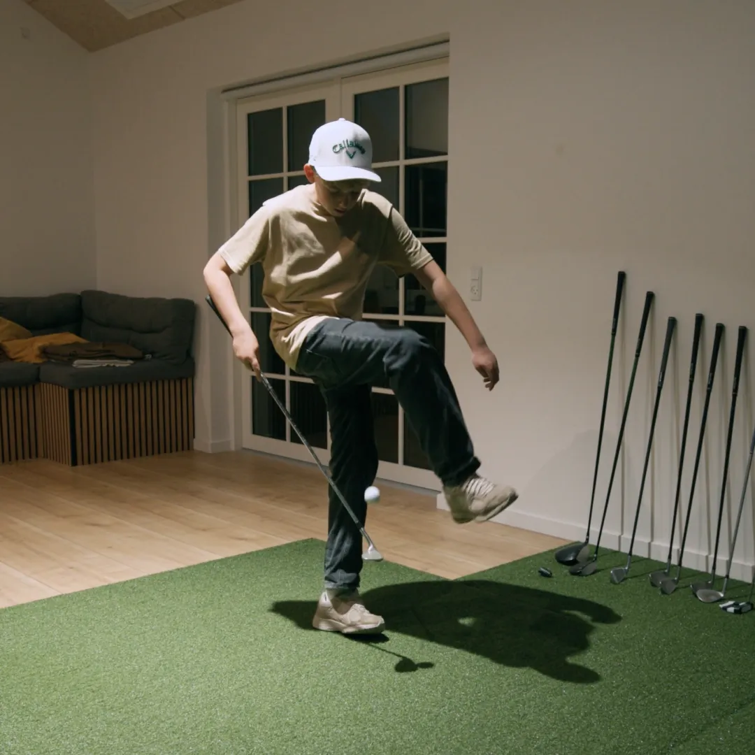 A person wearing a cap performs a trick with a golf club and ball inside a room, standing on green carpet. Golf clubs are lined up nearby.