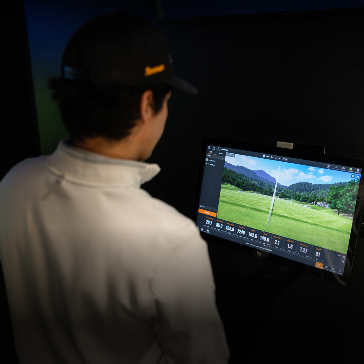 Person in a cap and jacket analyzes golf swing data on a monitor displaying a golf course and performance metrics.