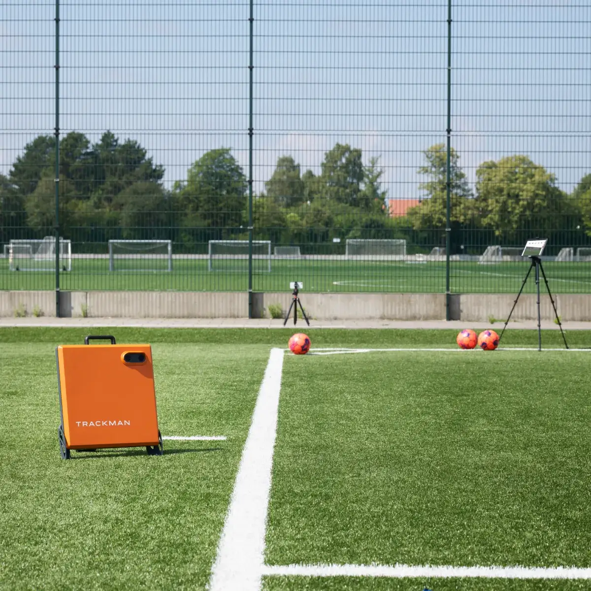 Soccer field with training equipment, including an orange Trackman device, cameras, and soccer balls on the grass near the goal area.