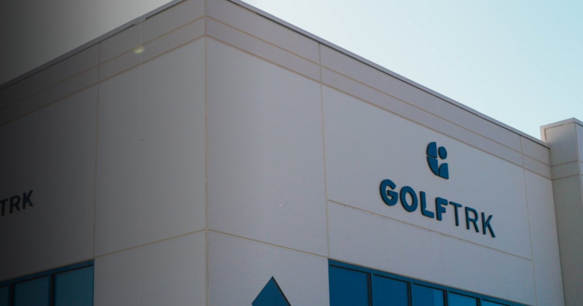 Exterior of a building with the "GOLFTRK" logo on the wall, featuring a geometric design and windows reflecting the clear sky.