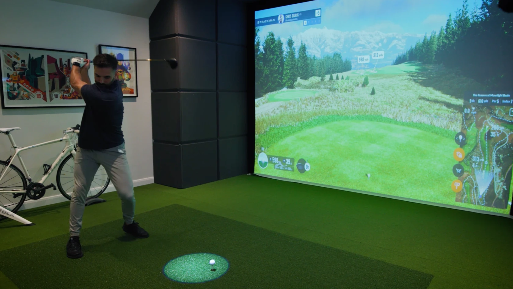 Person playing indoor golf simulator, swinging a club on a green mat. A screen displays a golf course scene. Bicycles and artwork in the room.