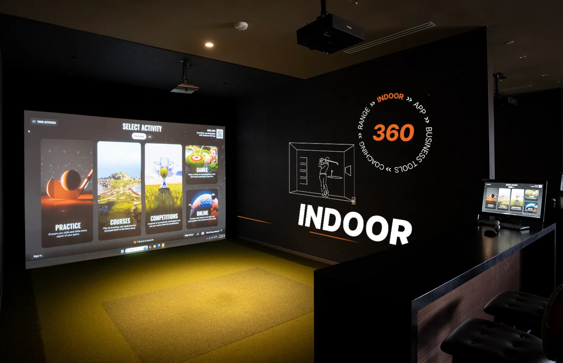 Indoor golf simulator setup with a large screen displaying various game modes and a side area with control panels and seating.