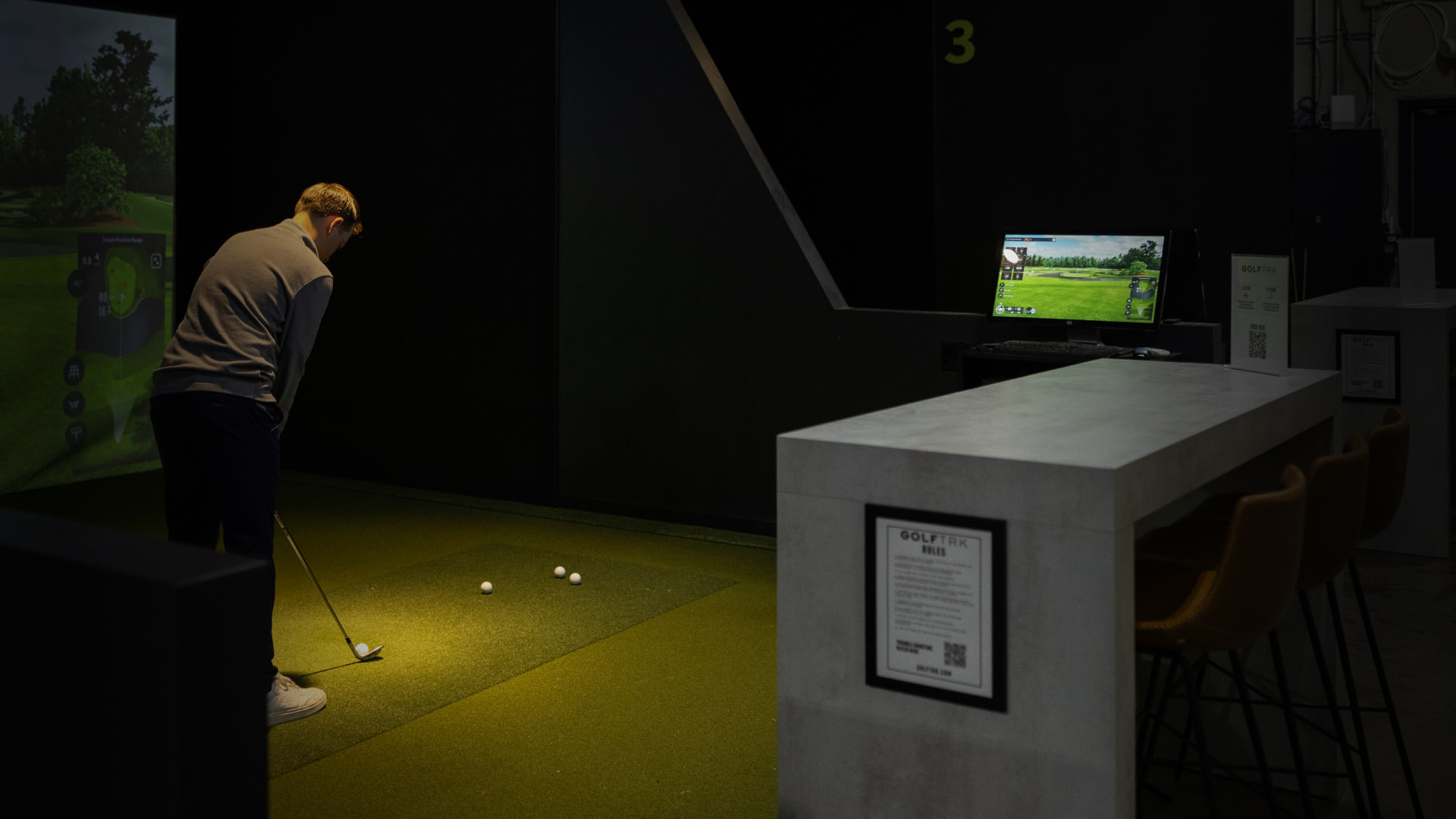 Person playing indoor golf with a simulator, putting on a green mat. A screen displays a golf course in the background.