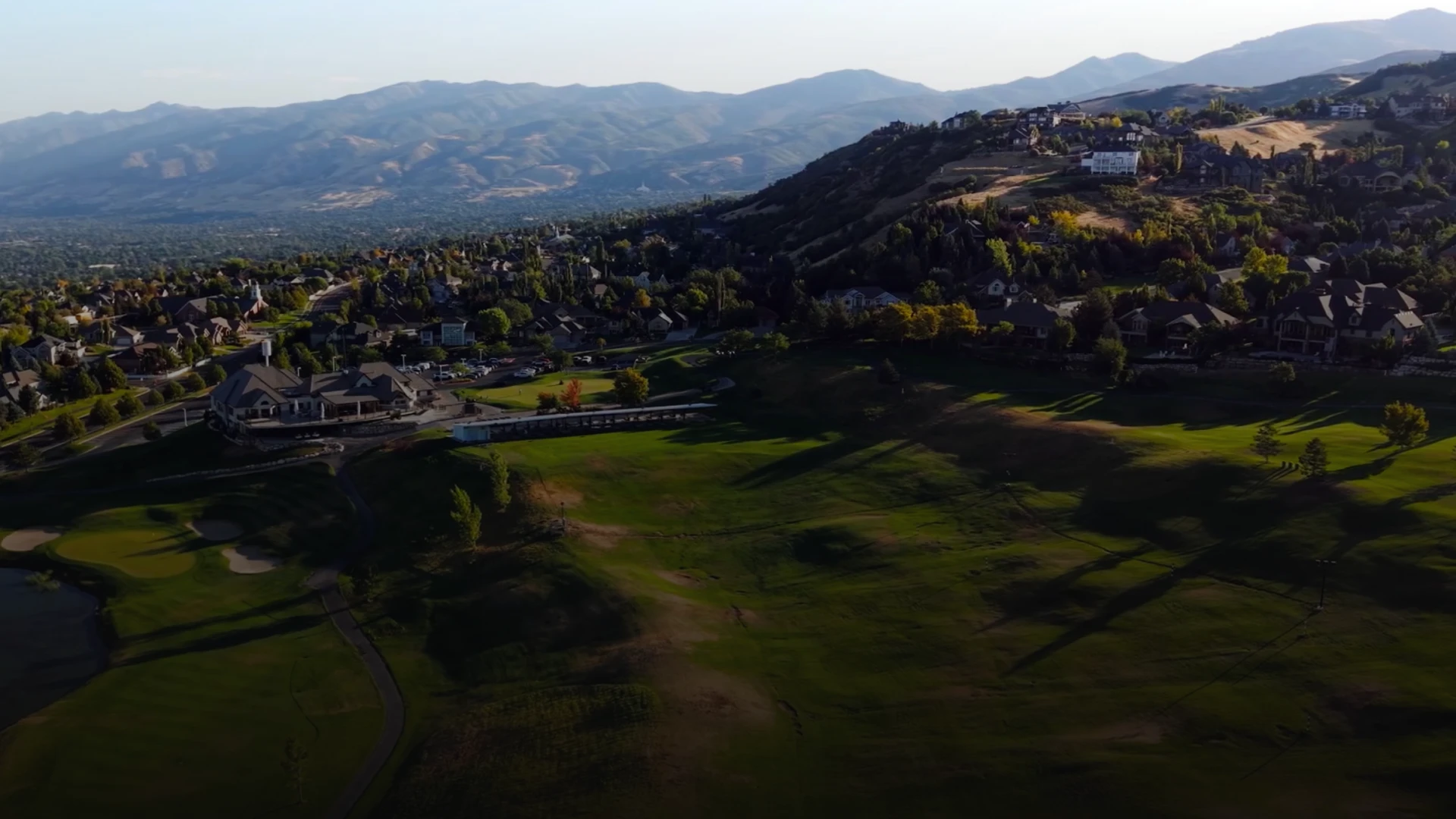 Aerial view of a hilly landscape with houses, a golf course, and distant mountains under a clear sky.