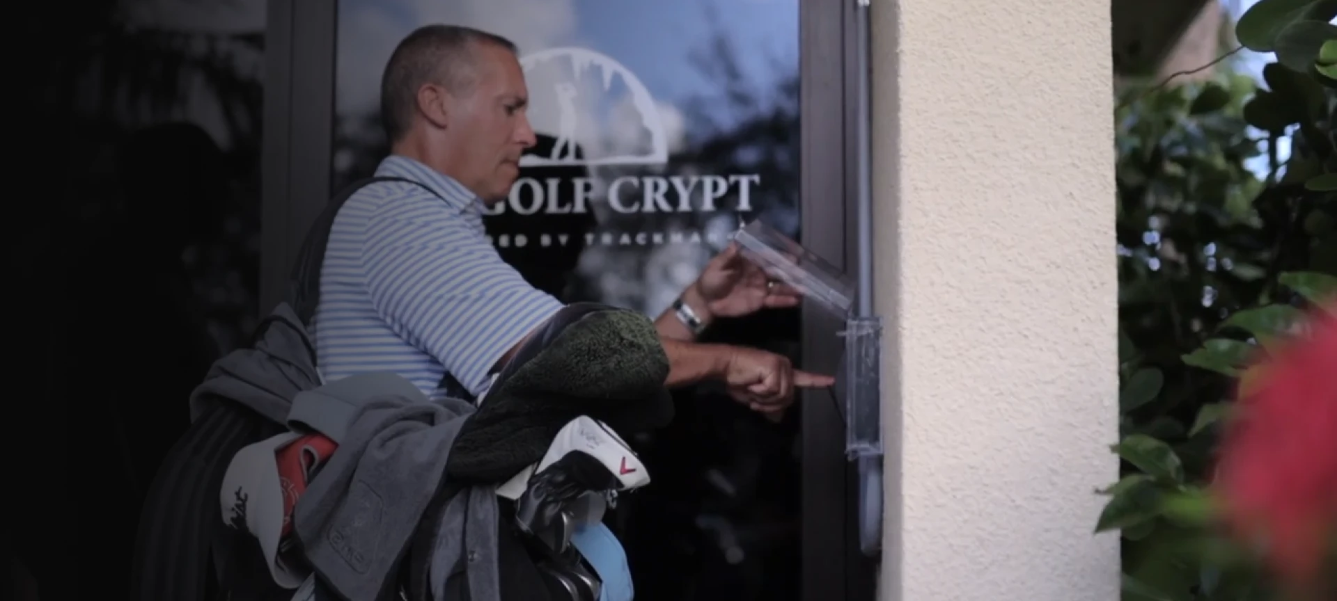 Man in striped polo carrying golf clubs taps access pad beside glass door labeled "GOLF CRYPT."