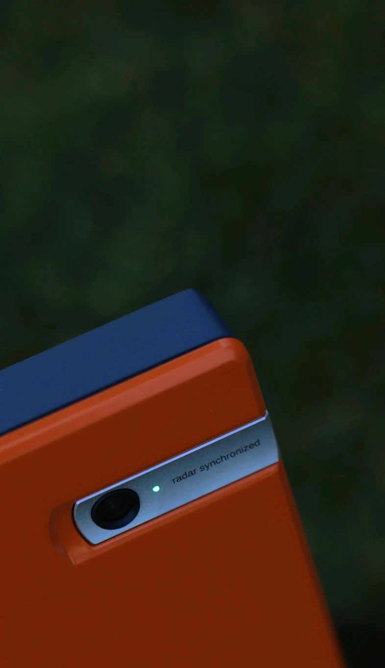 Close-up of an orange device with a camera sensor, labeled "radar synchronized," emitting a green light against a blurred dark background.