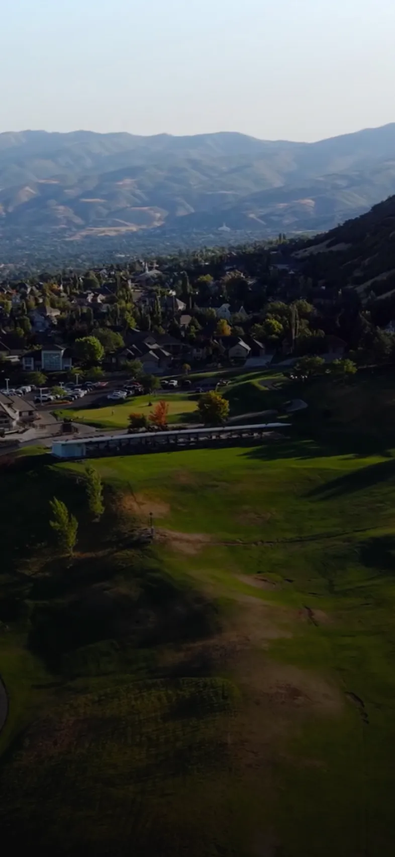 Aerial view of a suburban area with houses, a road, and a large grassy field surrounded by hills and mountains in the background.
