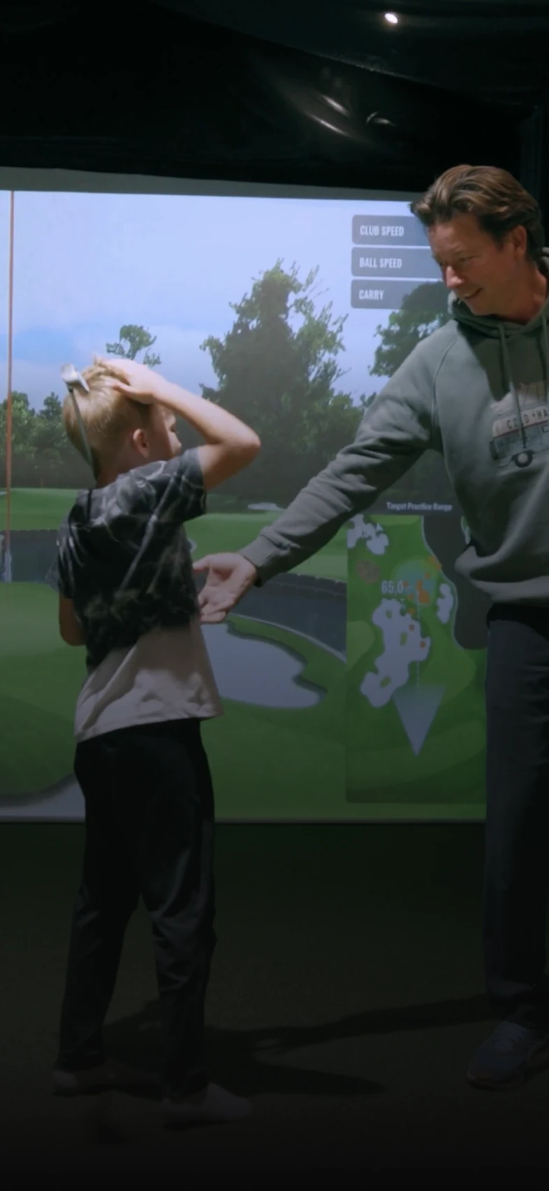 A person in a hoodie instructs a child on a golf swing in front of a golf simulator screen displaying course details.