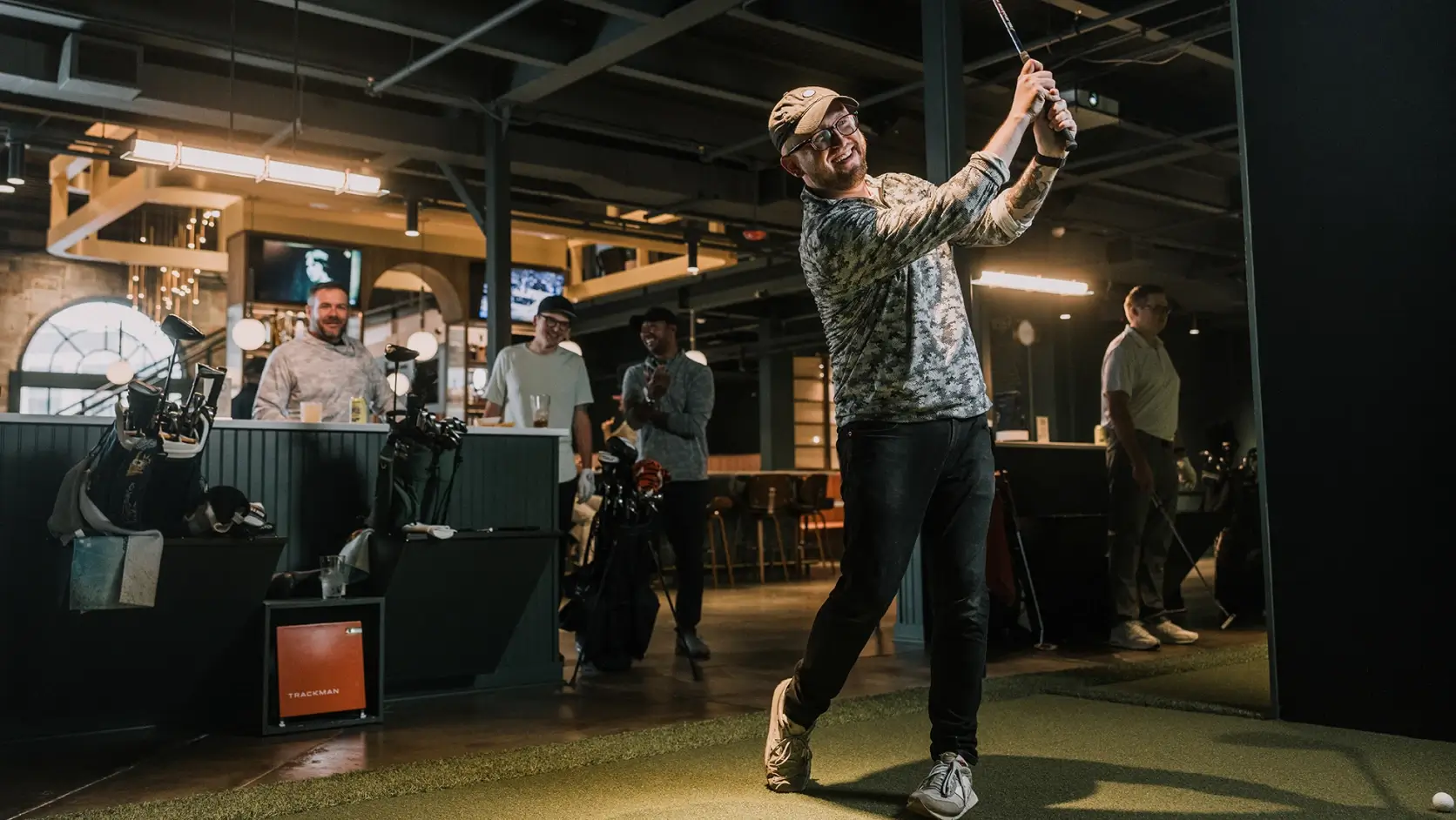 A man in a golf pose indoors, smiling, with onlookers and golf equipment in the background. Indoor setting with modern decor.
