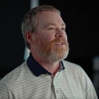 A bearded man in a striped polo shirt looks to the side against a dark background.