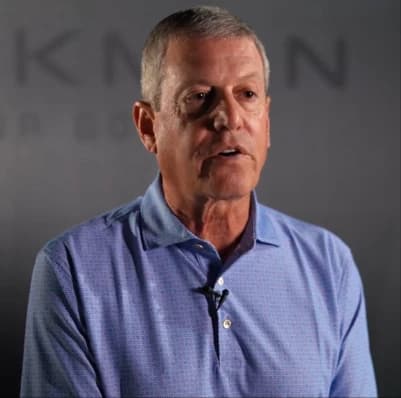 Middle-aged man in a light blue polo shirt speaking on camera with a lapel microphone against a dark blurred backdrop.