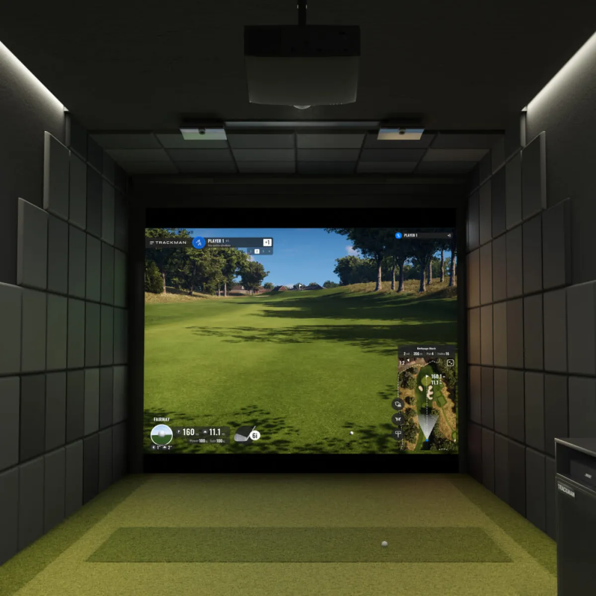 Indoor golf simulator with a large screen displaying a golf course, surrounded by padded walls and ceiling, with a golf ball on the floor.