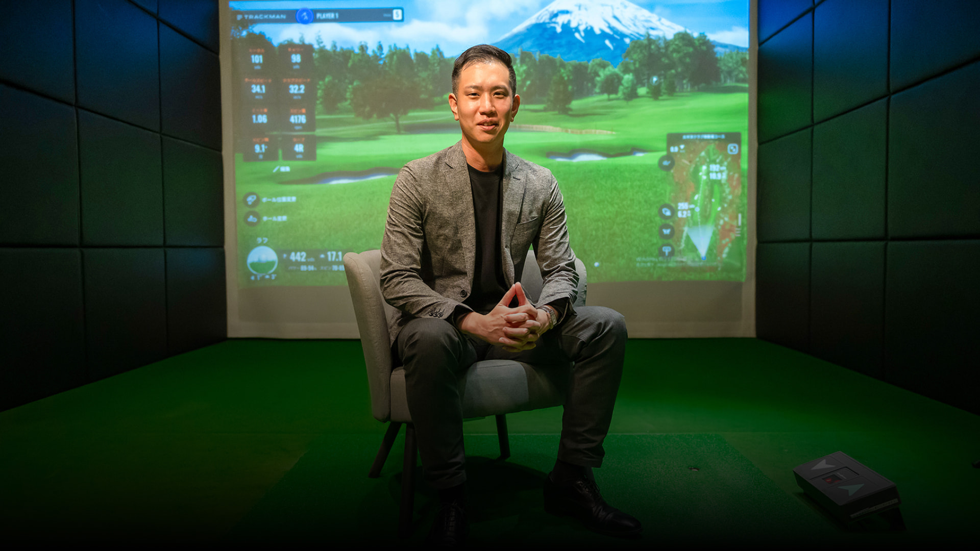 Man sitting on a chair in a golf simulator room, with a virtual golf course displayed on the screen behind him.