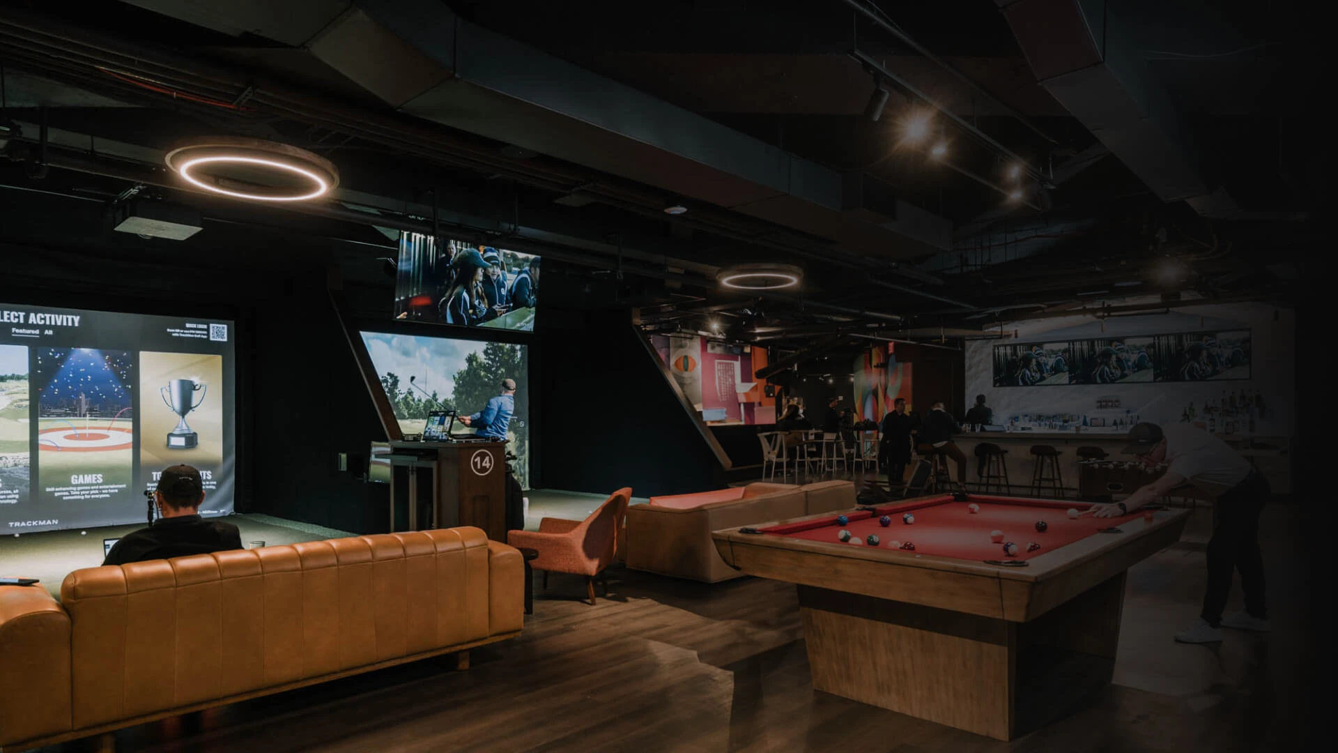 A dimly lit lounge with pool tables, a bar area, and screens displaying games, creating a relaxed and modern atmosphere.