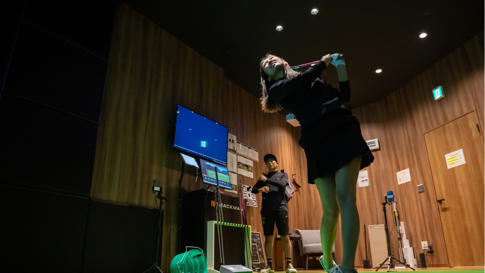 A woman swings a golf club indoors, while a man stands in the background. The room has wood-paneled walls and a screen displaying data.