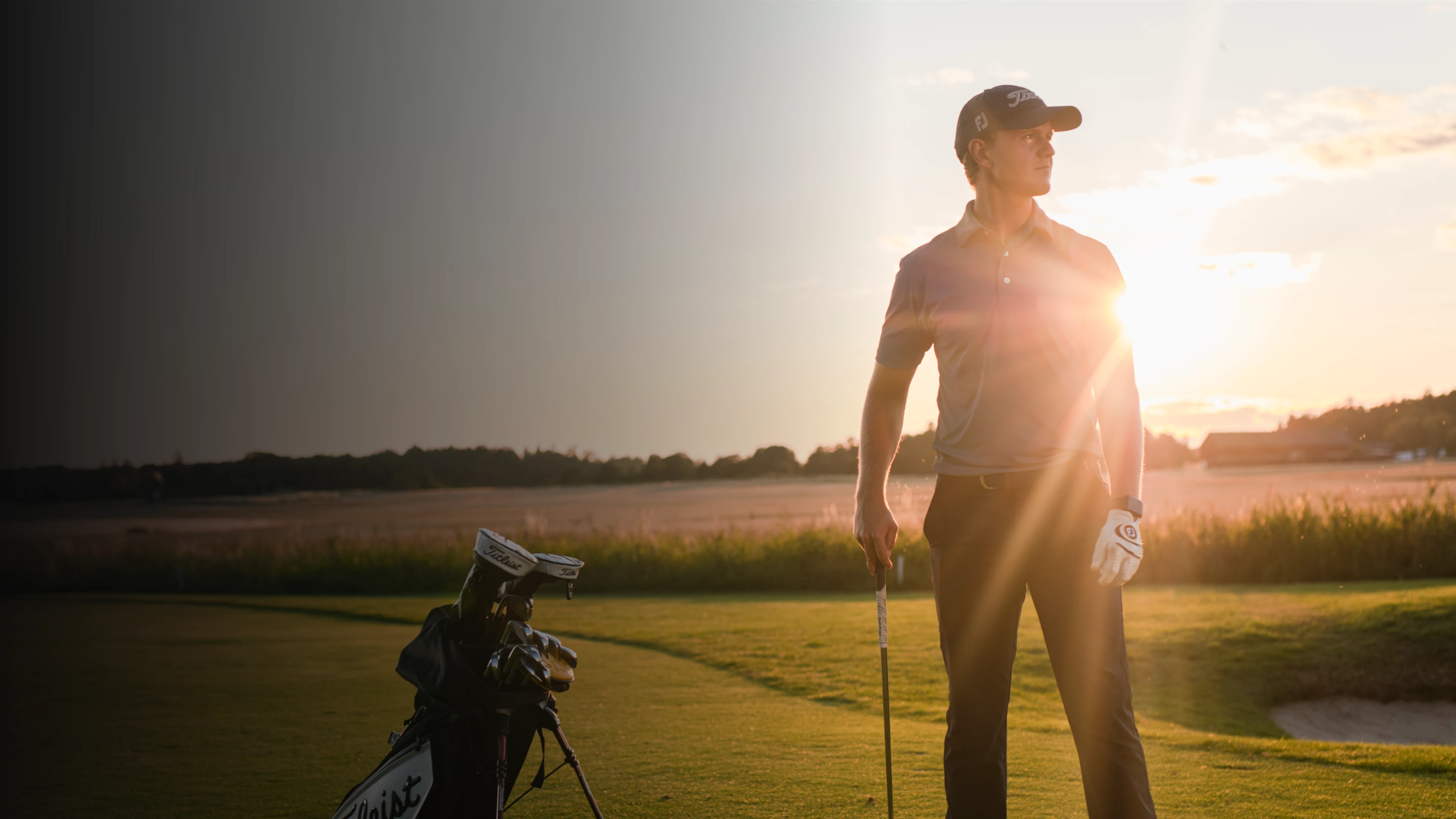 Golfer standing on a sunlit course holding a club, with a golf bag nearby. Sun is setting on the horizon, creating a warm glow.