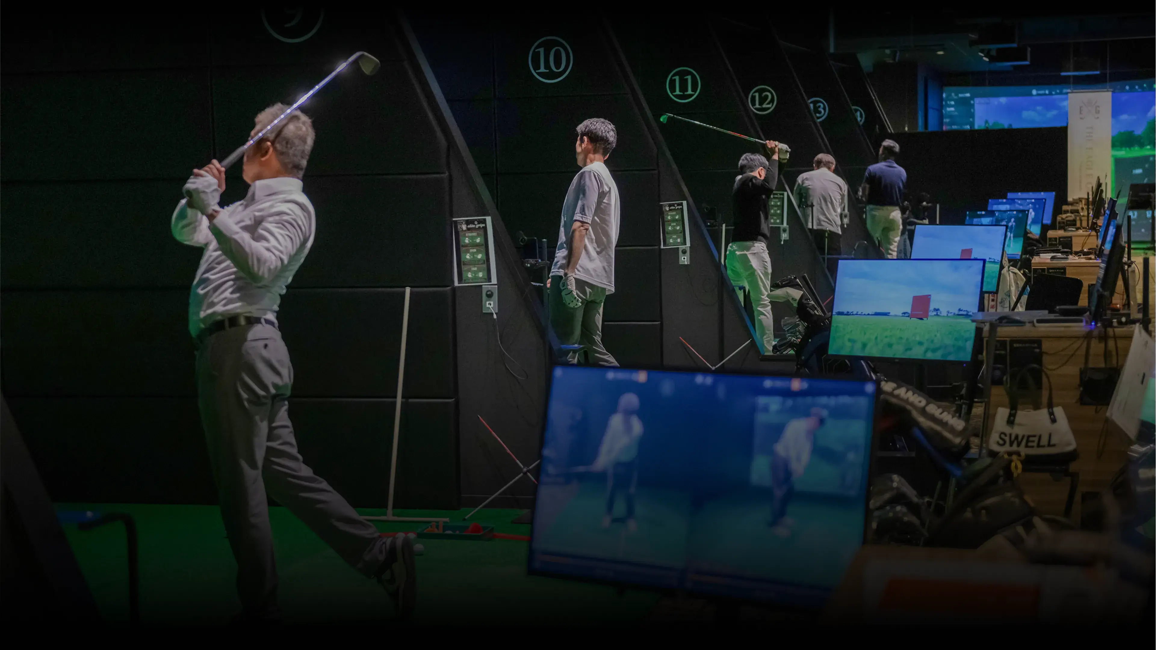 People practicing golf swings at an indoor golf range with numbered stalls and screens displaying swing analyses.