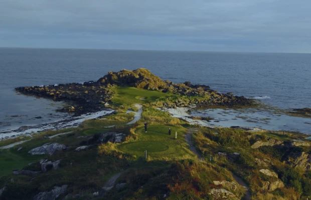 Aerial view of a coastal golf course with rocky terrain, surrounded by the ocean under a cloudy sky.