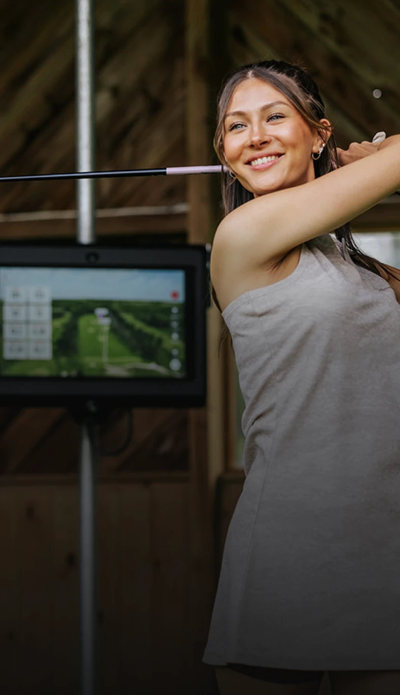 Woman smiling while swinging a golf club indoors, with a digital screen displaying a golf course in the background.