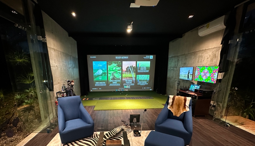 A Trackman home indoor golf simulator with a large screen displaying a golf game, two blue chairs, a zebra-patterned rug, and a small desk with monitors.